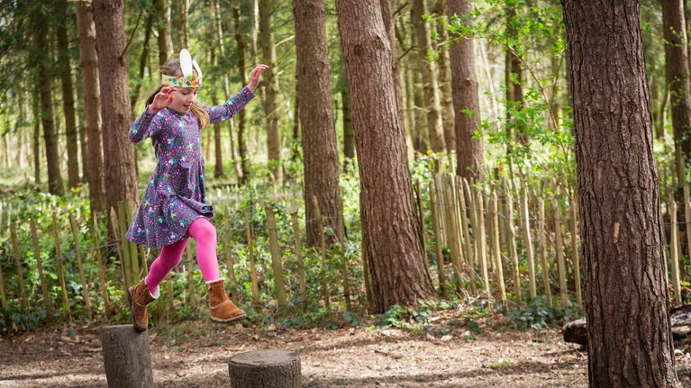 A child leaps across tree stumps in the natural play area at Calke Abbey, surrounded by green woodland and wearing Easter bunny ears.